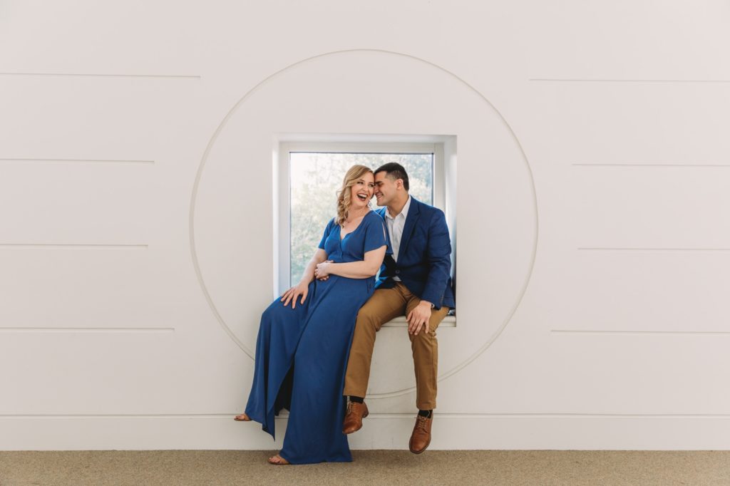 Engaged couple sitting in front of a white wall at Fernbank during their engagement session